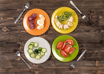 Four plates orange, yellow, green, white with sliced various foods and four forks on an old wooden table