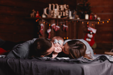 Fototapeta na wymiar cute young family on bed with Christmas background behind. Dad and mom kiss baby girl.