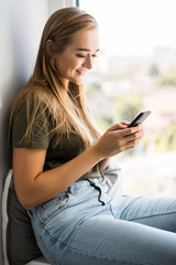 Fototapeta na wymiar Gorgeous young woman checking email and sending sms message on mobile phone sitting on windowsill