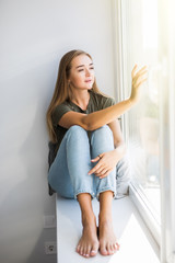 Young woman is opening window while sitting on windowsill