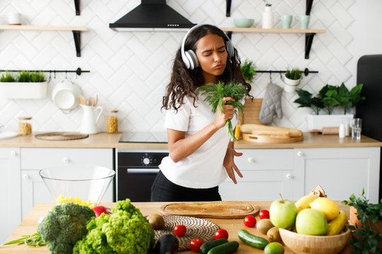Hilarious mulatto girl with closed eyes in big headphones is smiling and emotionally pretending like she is singing in greenery near the table with fresh vegetables and fruits