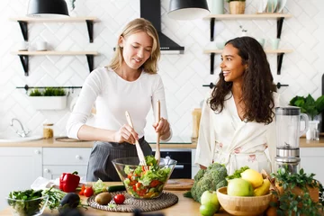Poster Attractive caucasian girl is cooking healthy salad and beautiful girl is looking  on her dressed in silky nightgown on modern designed kitchen © IVASHstudio