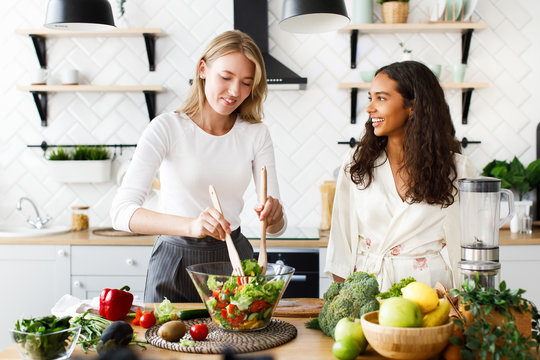 Attractive caucasian girl is cooking healthy salad and beautiful mulatto girl is looking  on her dressed in silky nightgown on modern designed kitchen