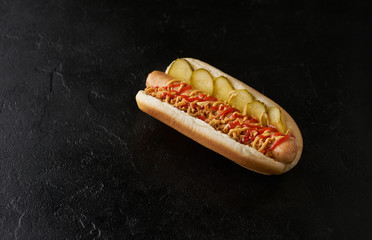 Hot dog with pickled cucumbers, fried onions, ketchup and mustard on a black background.
