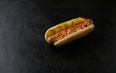 Hot dog with pickled cucumbers, fried onions, ketchup and mustard on a black background.