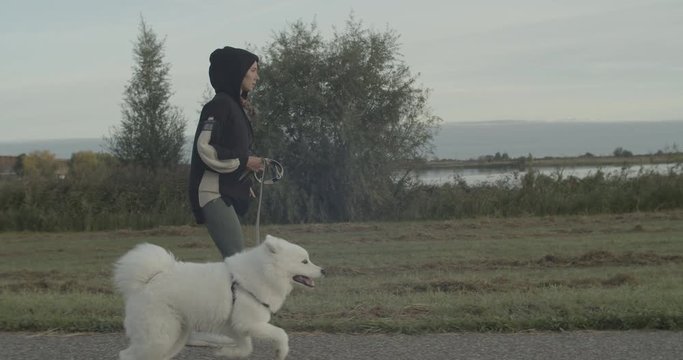 Young woman wearing black hoodie getting fun with her big white samoyed dog while running in the early morning near the river