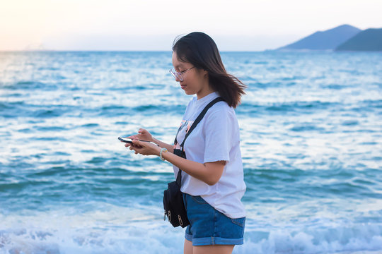A girl with an Asian appearance with a rose on a berug against the background of the sea, looks at a mobile phone. Young woman reads or writes messages on her smartphone