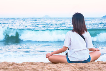 Fototapeta na wymiar A young woman sits in a lotus position on the shore on the sand, enjoys the sea or ocean.Cute Asian woman meditates and relaxes on the beach, in the evening at sunset, soft focus. copy space.