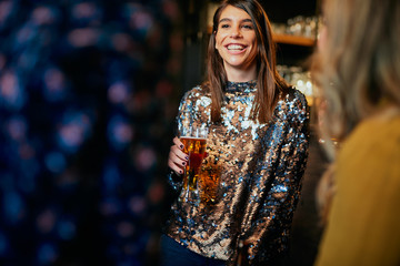 Fototapeta premium Beautiful caucasian smiling stylish woman holding beer and chatting with friends. In background is bar counter. Pub interior.