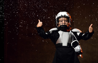 Boy in hockey uniform, helmet with skates on neck standing showing good thumb sign with fingers over dark background with snow