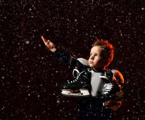 Fototapeta na wymiar Serious boy in protective hockey uniform with skates on neck standing and reaching up over dark background with snowflakes