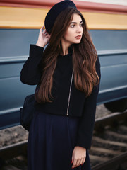 Obraz premium Young beautiful brown haired woman in black jacket blue skirt with gorgeous long legs in vintage beret emotionally posing on railway station train background with cinematic color grading