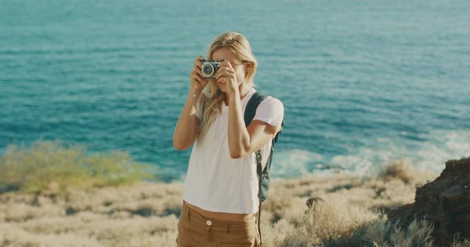 Attractive young blonde woman smiling and taking pictures with her vintage camera on a golden coast with ocean in the background, outdoor travel photographer woman taking photographs on vacation