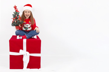 A little girl in a Santa hat sits on a Red cube in the Studio with a Christmas tree in her hands, on a white background. The concept of a happy childhood. Blank space for text. Layout for design