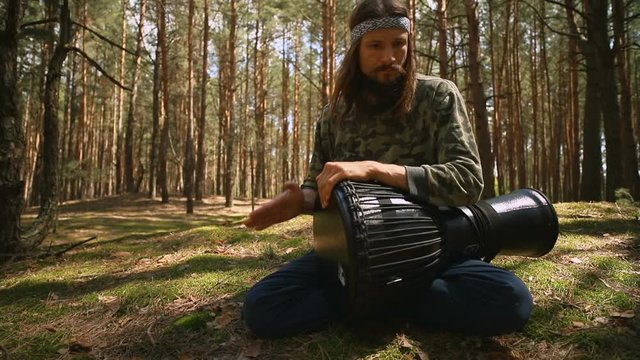 Young Caucasian hippie man playing a djembe drum in the summer coniferous forest