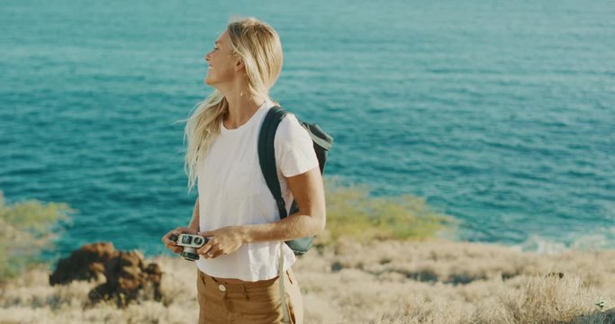 Attractive young blonde woman smiling and taking pictures with her vintage camera on a golden coast with ocean in the background, outdoor travel photographer woman taking photographs on vacation