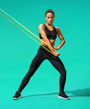 Athletic girl performs exercises using resistance band. Photo of young latin girl pumping biceps on turquoise background. Strength and motivation