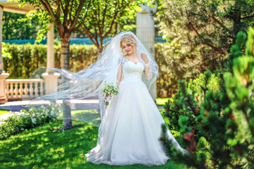 Obraz na płótnie Canvas Young bride wearing gorgeous wedding dress, playing with veil. plays a wedding dress. Dress develops in the wind. Happy blonde bride in park