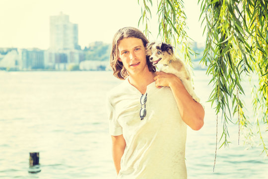 Portrait of man with best friend - dog. Young American Man with long hair, wearing white shirt, carrying dog on shoulder, standing by Hudson River with green willow leaves in New York City, relaxing..