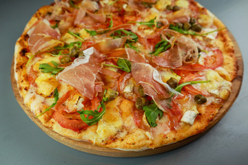 The pizza is prosciutto. Capers, tomatoes, arugula, mozzarella, brie cheese. Wooden plate. Gray background. Restaurant