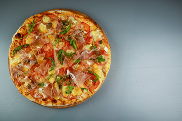 The pizza is prosciutto. Capers, tomatoes, arugula, mozzarella, brie cheese. Wooden plate. Gray background. Restaurant