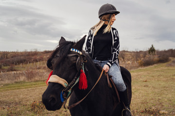 Young beautiful blonde caucasian woman riding a horse horseback casual rider girl in autumn or winter day wearing protective helmet