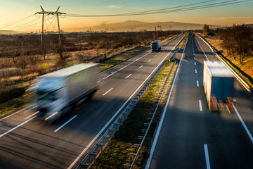 Delivery trucks in high speed driving on a highway through rural landscape. Fast blurred motion drive on the freeway. Freight scene on the motorway