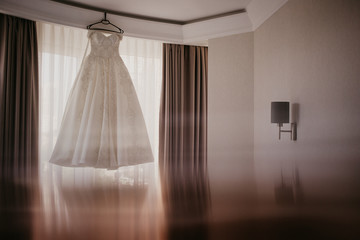 Wedding dress on hanger near window in the bride room. On her wedding day. Reflection on the bottom