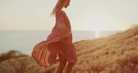 Happy carefree beautiful woman twirling her pink dress in the wind at sunset in a golden field by the sea, beautiful natural fashion model dancing at sunset - Powered by Adobe