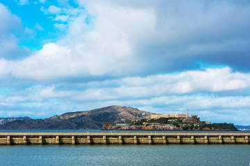 Obraz premium Scenic view of Alcatraz and Angel islands from the harbor safeguarded by concrete breakwater wall on the cloudy day