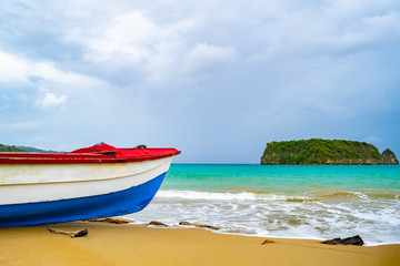 Fototapeta premium Colorful old wooden fishing boat docked by water on a beautiful beach coast land. White sand sea shore landscape on tropical Caribbean island. Holiday weekend/ summer vacation setting in Jamaica.