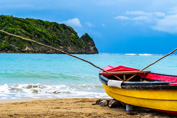 Naklejka na ściany i meble Colorful old wooden fishing boat docked by water on a beautiful beach coast. White sand sea shore landscape on tropical Caribbean island. Holiday weekend summer vacation setting in Jamaica.