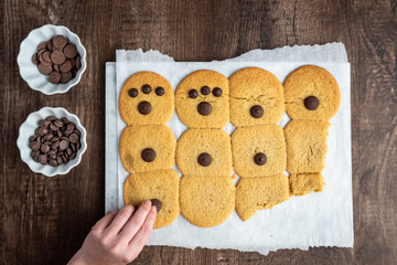 Two bowls of chocolate chips and batch of fresh cooked sugar cookies on parchment paper, woman’s hand putting chocolate on cookies
