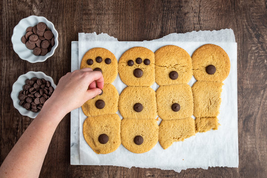 Two bowls of chocolate chips and batch of fresh cooked sugar cookies on parchment paper, woman’s hand putting chocolate on cookies