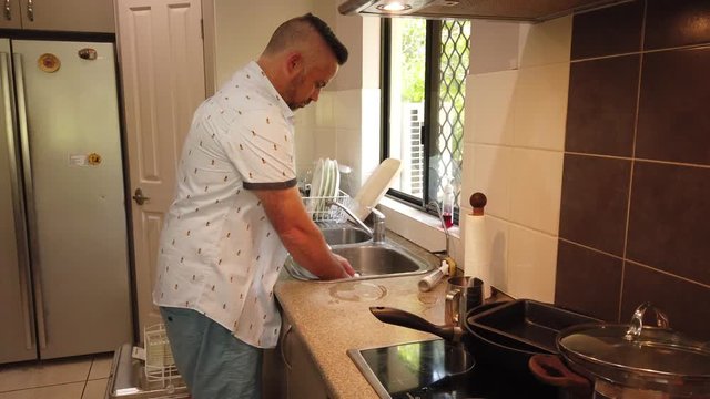 man in the kitchen washing a decorative coffee mug in the sink in front of the window. Light coming in from the window, looking from over the stove top, rinsing and placing the mug in the dryer,