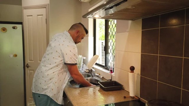 Man at a sink in front of a window with natural light washing a large frying pan in the sink, rinsing the pot and placing it on the stove to dry