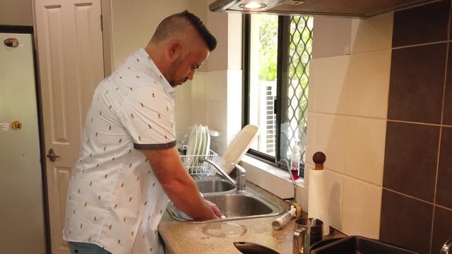 man doing the dishes in a well lit kitchen with the sun coming through the window, washing a brown coffee mug, rinsing and placing in the dryer