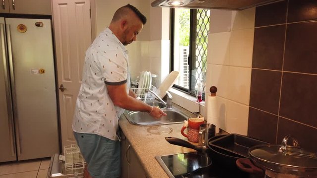 guy in the kitchen cleaning dishes at the sink by the window, cleaning the cutlery one piece at a time and rinsing it then putting it in the dish rack