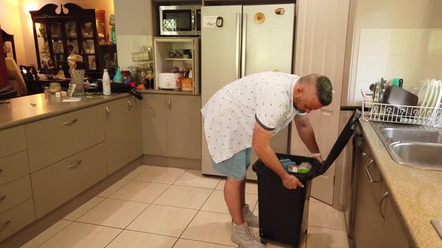 man in casual clothing emptying the kitchen garbage bin to put a bew bin liner in it,