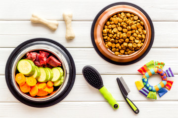 Pet care concept. Dry feed and meat in bowls near toy, brush and bones on white wooden background top view
