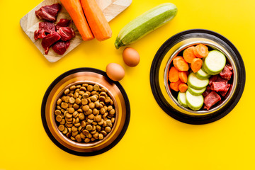 Pet feed ingredients. Raw meat and fresh vegetables near bowl with dry feed on yellow background top-down