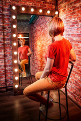 Girl in red shirt on a tall chair in front of a lighted mirror in a dark room. Model posing for a...
