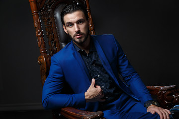 Elegant man in a classic blue suit sitting in a carved wooden chair against a dark background.