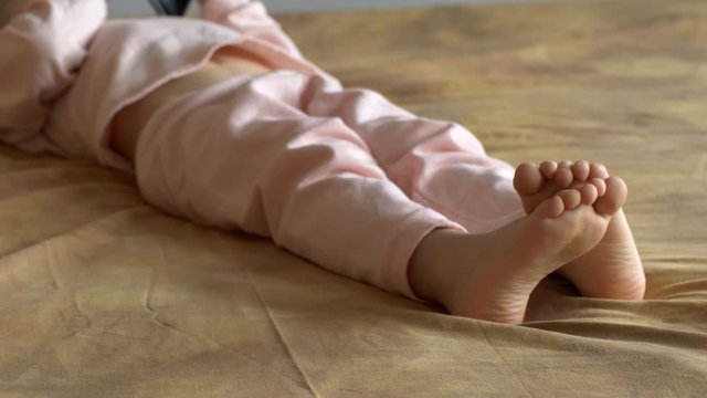 Legs, feet and fingers of boy close-up. Cute blond child in pink pajamas plays with touch screen of smartphone while lying on sofa. the kid wants learn everything. Home teaching for lagging children
