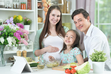 Happy pregnant woman with husband and daughter preparing salad together at kitchen