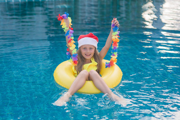 Kid wearing Santa Claus hat are swimming in a blue pool on a bright sunny day and smiling. Concept of happy new year and christmas.