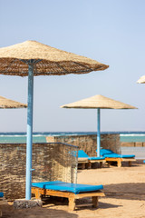 Wooden sun beds by the sea covered with blue towels and with wickered sunshades