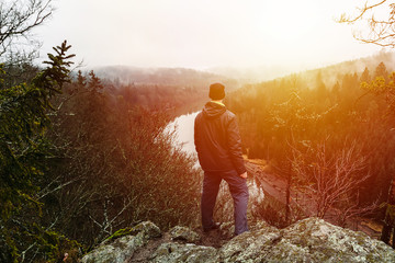 Young man in jacket, pants and backpack standing on rock looking to river Vltava and valley at sunset.