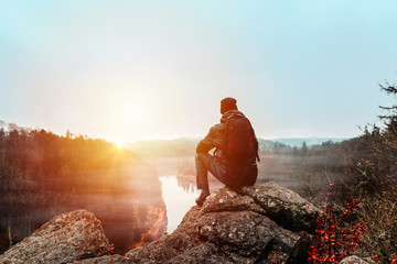 Young man in jacket, pants and backpack sitting on rock looking to river Vltava and valley in...