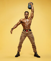 Fototapeta na wymiar Young muscular guy training with kettlebell. Photo of handsome man with naked torso on yellow background. Strength and motivation. Full length